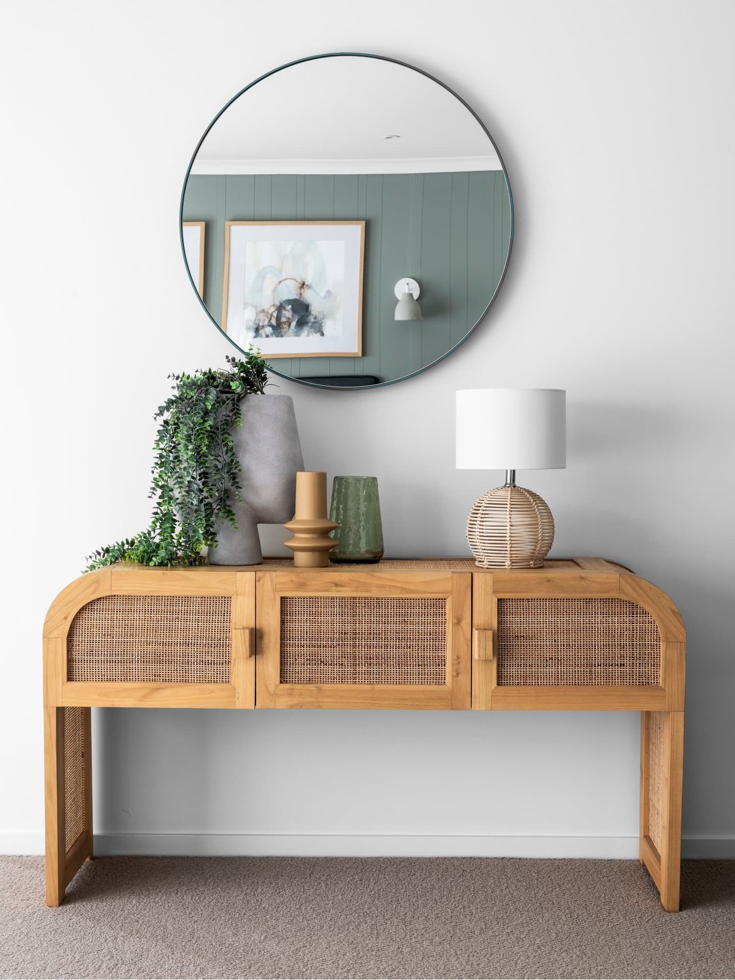 rattan curved console table with round mirror above styled with coastal table lamp and concrete vase
